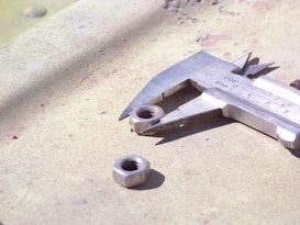 A close-up view of a metallic caliper measuring the diameter of a hexagonal nut. The caliper is placed on a textured surface, accompanied by another nut nearby. The lighting creates a shiny effect on the metal parts, highlighting their contours and precision.