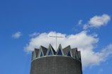A modern church tower topped with a cross against a backdrop of a clear blue sky with a few fluffy white clouds. The tower has a dark, conical roof featuring triangular windows that resemble a crown.