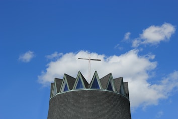 A modern church tower topped with a cross against a backdrop of a clear blue sky with a few fluffy white clouds. The tower has a dark, conical roof featuring triangular windows that resemble a crown.