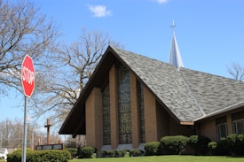 A large church building with a steep, gabled roof and patterned windows stands against a clear blue sky. In front of the church is a stop sign and some bare trees. The church features a tall steeple topped with a cross.