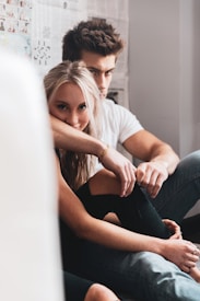 Two people sit closely together by a wall covered in newspaper. One person, with long blonde hair, is leaning slightly forward with their arm resting on their knee, looking directly at the camera. The other person is partially behind them, with short brown hair, wearing a white t-shirt, and gazing intently sideways.