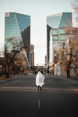 A person wearing a ghost costume made from a white sheet stands in the middle of an empty city street. Tall modern buildings with reflective glass surfaces line the street, and bare trees are visible along the sidewalks.