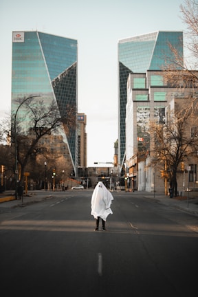 A person wearing a ghost costume made from a white sheet stands in the middle of an empty city street. Tall modern buildings with reflective glass surfaces line the street, and bare trees are visible along the sidewalks.