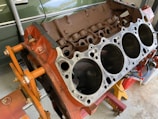 A disassembled car engine block with visible cylinders, mounted on an orange engine stand. The background includes a green vehicle partially visible and some tools.