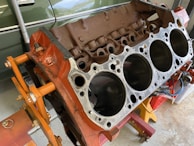 A disassembled car engine block with visible cylinders, mounted on an orange engine stand. The background includes a green vehicle partially visible and some tools.