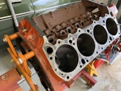 A disassembled car engine block with visible cylinders, mounted on an orange engine stand. The background includes a green vehicle partially visible and some tools.