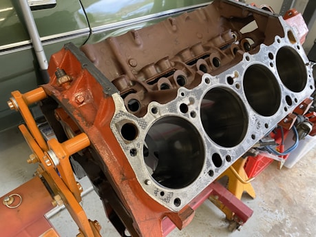 A disassembled car engine block with visible cylinders, mounted on an orange engine stand. The background includes a green vehicle partially visible and some tools.