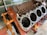 A disassembled car engine block with visible cylinders, mounted on an orange engine stand. The background includes a green vehicle partially visible and some tools.