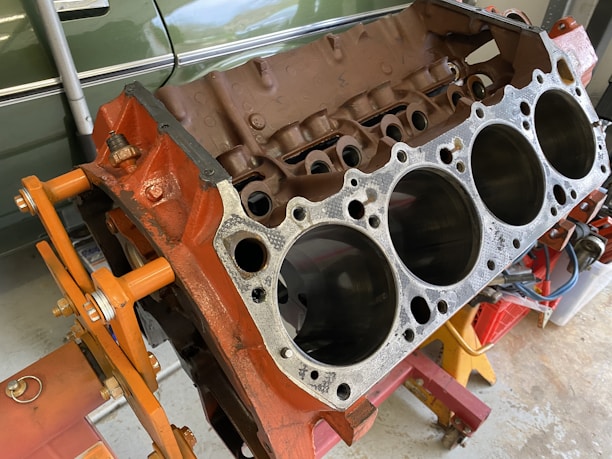 A disassembled car engine block with visible cylinders, mounted on an orange engine stand. The background includes a green vehicle partially visible and some tools.