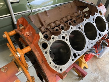 A disassembled car engine block with visible cylinders, mounted on an orange engine stand. The background includes a green vehicle partially visible and some tools.