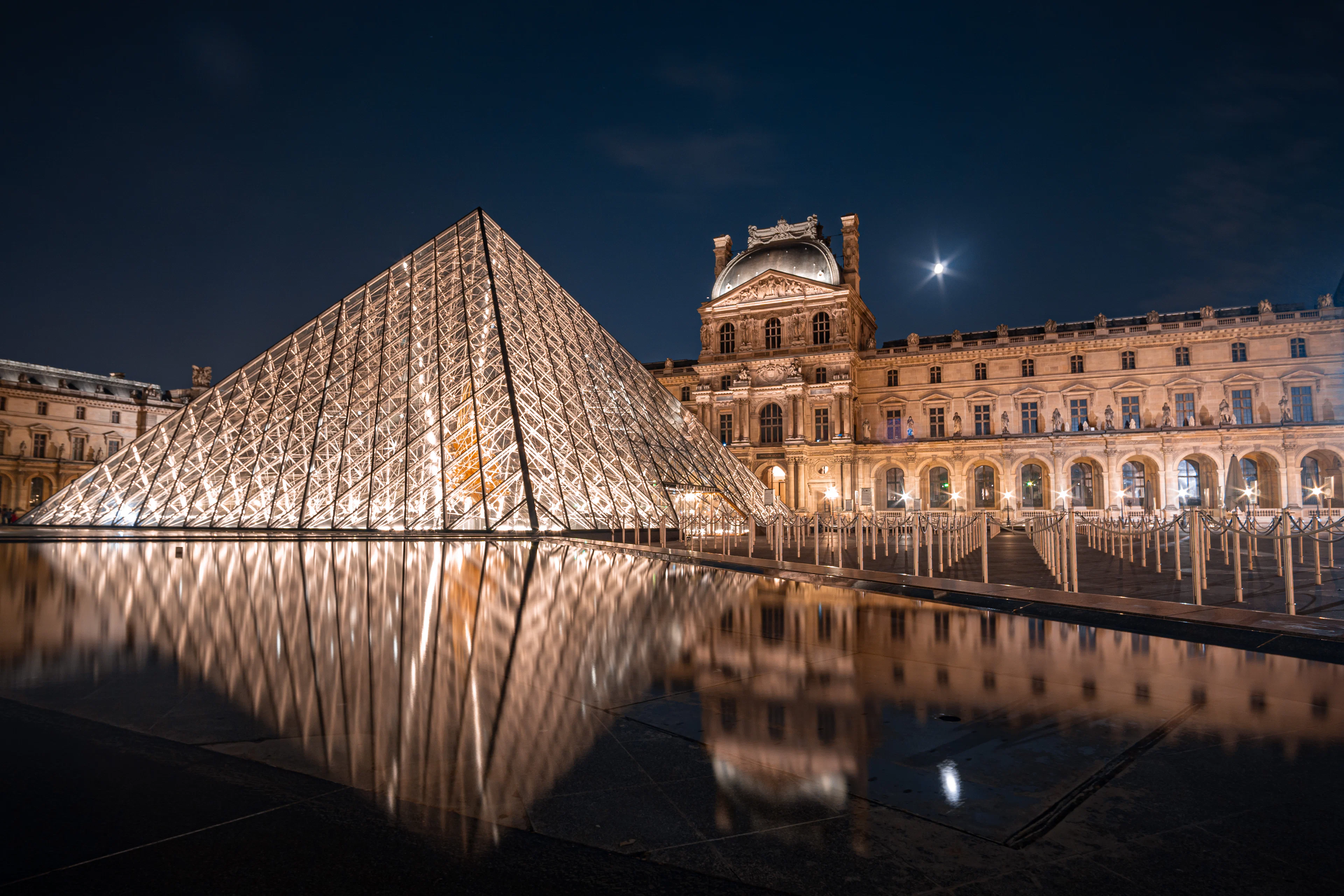 The illuminated Louvre Pyramid reflected in wet paving at night, with the Pavillon Sully and the moon