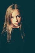 A young girl with long blonde hair and blue eyes is looking directly at the camera. The lighting casts a soft glow on her face, creating gentle shadows. Her expression appears contemplative or reflective, set against a dark background.