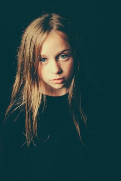A young girl with long blonde hair and blue eyes is looking directly at the camera. The lighting casts a soft glow on her face, creating gentle shadows. Her expression appears contemplative or reflective, set against a dark background.