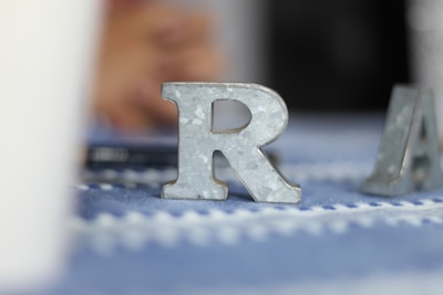A metallic letter 'R' stands upright on a patterned blue and white surface, with another metallic letter partially visible in the background. The surface has a soft focus, and the image has a calm ambiance.