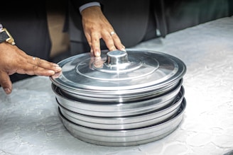 Two hands are seen resting on a stack of covered metal trays placed on a white surface. The hands appear to be those of adults, and the rings suggest they might be of an important event or occasion. The trays are shiny and metallic, reflecting light.