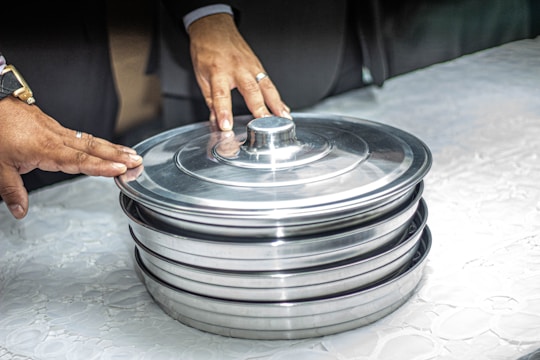 Two hands are seen resting on a stack of covered metal trays placed on a white surface. The hands appear to be those of adults, and the rings suggest they might be of an important event or occasion. The trays are shiny and metallic, reflecting light.