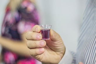 A hand holding a small, clear plastic cup filled with a dark red or purple liquid. In the blurred background, there is a person wearing a patterned clothing.