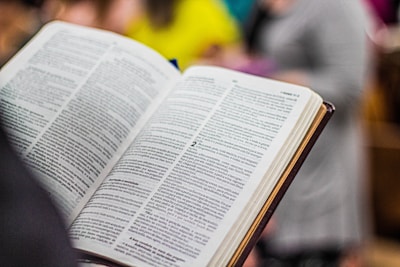 An open book with text in a foreign language is held by a person. The pages are filled with paragraphs, and the book has a hardcover with a light brown edge. In the background, the image is slightly blurred, showing indistinct colorful shapes suggesting people or objects.