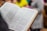An open book with text in a foreign language is held by a person. The pages are filled with paragraphs, and the book has a hardcover with a light brown edge. In the background, the image is slightly blurred, showing indistinct colorful shapes suggesting people or objects.