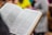 An open book with text in a foreign language is held by a person. The pages are filled with paragraphs, and the book has a hardcover with a light brown edge. In the background, the image is slightly blurred, showing indistinct colorful shapes suggesting people or objects.