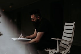 A man sits on a white chair in a dimly lit environment, writing in a notebook with focused attention. The surroundings are dark, with a beam of light illuminating the notepad and his hands.