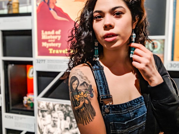 A person with curly dark hair is wearing denim overalls and beaded earrings. They are showing off a detailed tattoo on their upper arm of a woman holding flowers. The background includes various vibrant posters and images, adding an eclectic and artistic atmosphere.