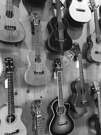 Various guitars and ukuleles are displayed hanging vertically on a wooden wall, arranged in a compact manner. They vary in shapes and sizes, featuring different designs and materials. The image is presented in black and white.