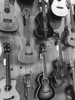 Various guitars and ukuleles are displayed hanging vertically on a wooden wall, arranged in a compact manner. They vary in shapes and sizes, featuring different designs and materials. The image is presented in black and white.