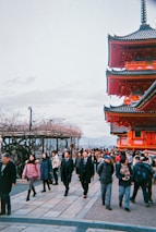 A busy scene featuring a mix of tourists and locals walking along a path. On the right, a traditional red Japanese temple or pagoda stands prominently with intricate architectural details. The sky is overcast, and there are bare trees on the left side, indicating a possible winter season.