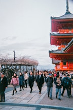 A busy scene featuring a mix of tourists and locals walking along a path. On the right, a traditional red Japanese temple or pagoda stands prominently with intricate architectural details. The sky is overcast, and there are bare trees on the left side, indicating a possible winter season.