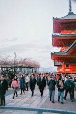 A busy scene featuring a mix of tourists and locals walking along a path. On the right, a traditional red Japanese temple or pagoda stands prominently with intricate architectural details. The sky is overcast, and there are bare trees on the left side, indicating a possible winter season.
