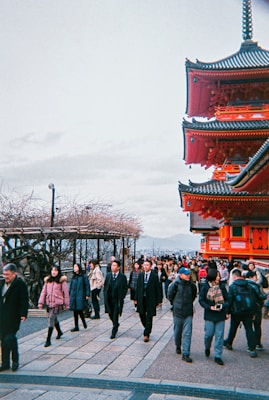 A busy scene featuring a mix of tourists and locals walking along a path. On the right, a traditional red Japanese temple or pagoda stands prominently with intricate architectural details. The sky is overcast, and there are bare trees on the left side, indicating a possible winter season.