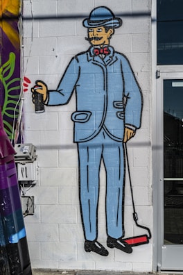 A mural of a man in a blue suit and bowler hat holding a paint roller and a spray can. The style is cartoonish, featuring bold outlines and bright colors. The man has a curled mustache and a bow tie, painted on a white brick wall next to a utility box.