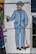 A mural of a man in a blue suit and bowler hat holding a paint roller and a spray can. The style is cartoonish, featuring bold outlines and bright colors. The man has a curled mustache and a bow tie, painted on a white brick wall next to a utility box.