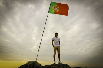 A person is standing on rocky terrain, looking off into the distance. A tall pole to the person's right, holds a large flag of Portugal, fluttering in the wind. Surrounding them is a dramatic sky with heavy, cloud cover.
