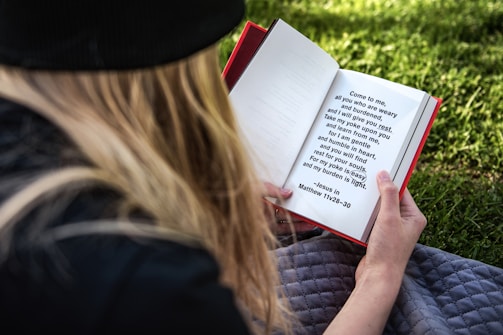 A person with long hair, wearing a black hat, is sitting on a grey blanket on a grassy area while reading a red-bound book. The text on the page is a Bible verse from Matthew 11:28-30, focusing on themes of rest and gentleness.