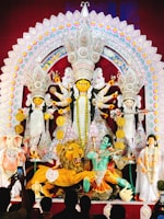 A grand and colorful depiction of a Hindu goddess surrounded by various symbolic figures and elements. The central figure is adorned with intricate ornaments and is holding multiple weapons, suggesting a scene of divine power. To the sides, there are figures including a lion, an elephant-headed figure, and others, all rich in traditional attire and symbolism. The backdrop is elaborately decorated with vibrant patterns and embellishments.