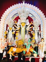 A grand and colorful depiction of a Hindu goddess surrounded by various symbolic figures and elements. The central figure is adorned with intricate ornaments and is holding multiple weapons, suggesting a scene of divine power. To the sides, there are figures including a lion, an elephant-headed figure, and others, all rich in traditional attire and symbolism. The backdrop is elaborately decorated with vibrant patterns and embellishments.