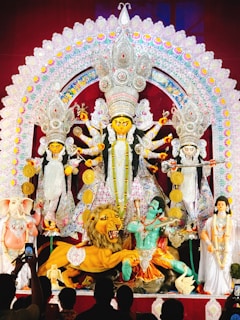 A grand and colorful depiction of a Hindu goddess surrounded by various symbolic figures and elements. The central figure is adorned with intricate ornaments and is holding multiple weapons, suggesting a scene of divine power. To the sides, there are figures including a lion, an elephant-headed figure, and others, all rich in traditional attire and symbolism. The backdrop is elaborately decorated with vibrant patterns and embellishments.