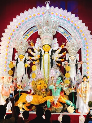 A grand and colorful depiction of a Hindu goddess surrounded by various symbolic figures and elements. The central figure is adorned with intricate ornaments and is holding multiple weapons, suggesting a scene of divine power. To the sides, there are figures including a lion, an elephant-headed figure, and others, all rich in traditional attire and symbolism. The backdrop is elaborately decorated with vibrant patterns and embellishments.