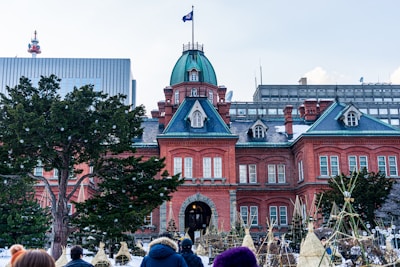 A large red brick building with a green dome and several windows. The building has a historical and European architectural style. In front, there are trees and several people standing or walking around. The sky is cloudy, and there are other modern buildings in the background.
