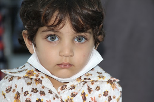 A young child with curly hair and striking blue eyes is wearing a floral-patterned shirt and a surgical mask pulled below their chin. The background is out of focus, highlighting the child's facial features and expressions.