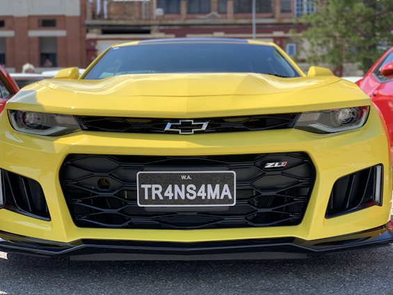 A yellow sports car with a prominent grille and sharp headlights is parked outdoors. The front license plate reads 'TR4NS4MA' and the car is shiny, reflecting the surrounding light. The Chevrolet logo is visible on the grille, and the ZL1 badge is displayed on the front.