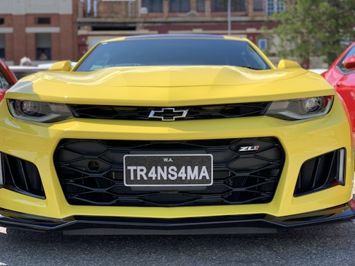 A yellow sports car with a prominent grille and sharp headlights is parked outdoors. The front license plate reads 'TR4NS4MA' and the car is shiny, reflecting the surrounding light. The Chevrolet logo is visible on the grille, and the ZL1 badge is displayed on the front.