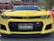 A yellow sports car with a prominent grille and sharp headlights is parked outdoors. The front license plate reads 'TR4NS4MA' and the car is shiny, reflecting the surrounding light. The Chevrolet logo is visible on the grille, and the ZL1 badge is displayed on the front.