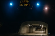 A concrete road tunnel with overhead lighting, visible under a dark sky. Traffic lights are mounted above the entrance, glowing blue, and an electronic sign displays a speed limit of 35 MPH.