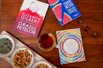 A wooden table with three books displayed alongside a cup of tea and a pair of glasses. One book features a colorful circular pattern on the cover, another has a vibrant pink and red design, while the third book shows a blue cover with illustrations of watermelon slices. A white tray with small bowls of almonds and seeds adds to the composition.