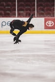 A person in a black ice skating outfit performs an athletic maneuver on an indoor ice rink. Their right arm is extended backwards, and their knees are bent, suggesting motion and control. The rink is empty, with maroon seats visible in the background. Advertisements line the boards around the rink.