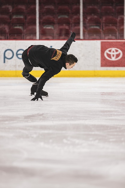 A person in a black ice skating outfit performs an athletic maneuver on an indoor ice rink. Their right arm is extended backwards, and their knees are bent, suggesting motion and control. The rink is empty, with maroon seats visible in the background. Advertisements line the boards around the rink.