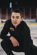 A person kneels on an ice rink wearing black athletic attire with 'Denver' and 'figure skating' embroidered on the jacket. The background shows an empty arena with blurred seats and advertisements.